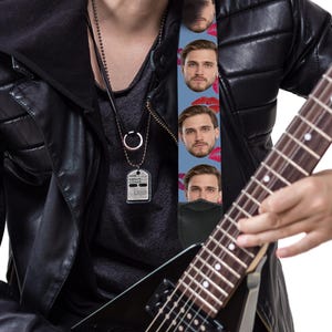 May include: A black electric guitar with a custom guitar strap featuring a repeating pattern of a man's face and red lips on a light blue background. The guitar strap is attached to the guitar. The person is wearing a black leather jacket.