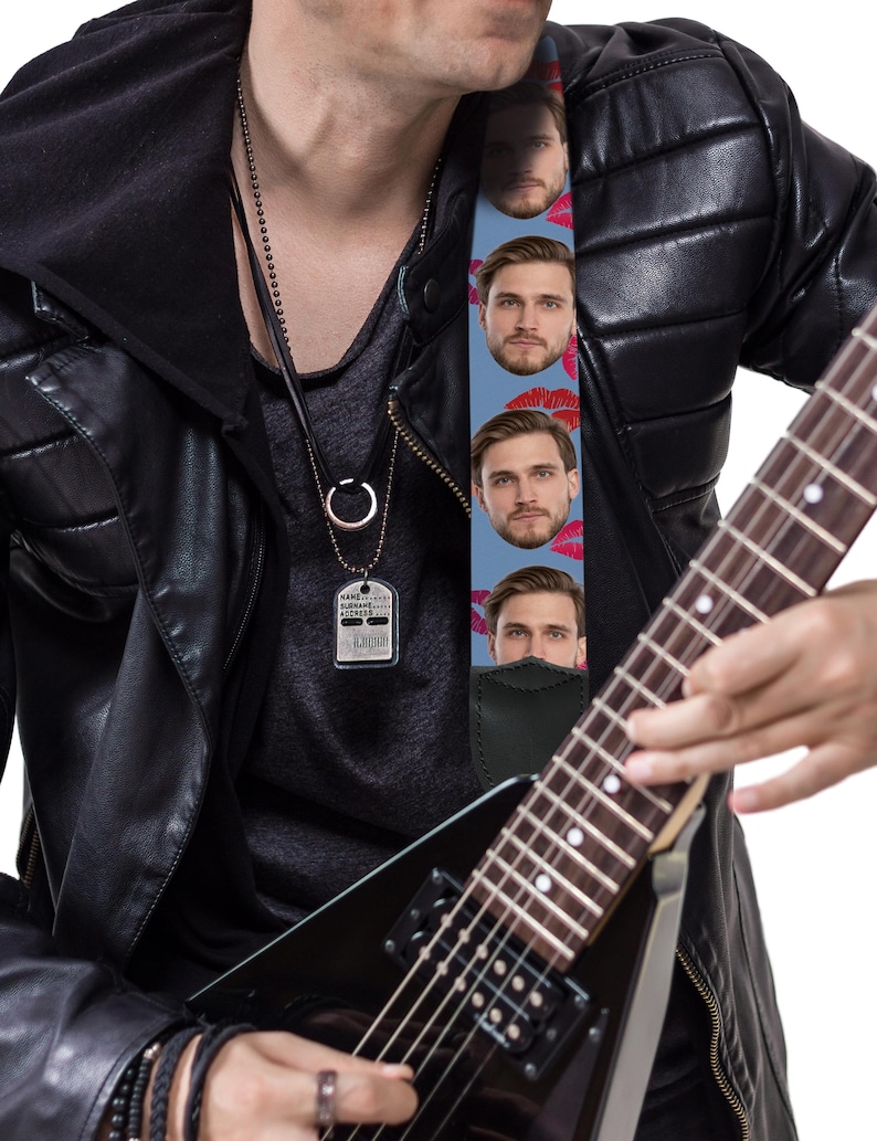 May include: A black electric guitar with a custom guitar strap featuring a repeating pattern of a man's face and red lips on a light blue background. The guitar strap is attached to the guitar. The person is wearing a black leather jacket.