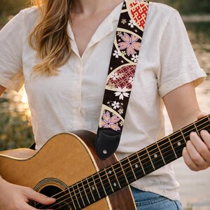 May include: A guitar strap with a black background and a Japanese-inspired floral design. The strap features pink cherry blossoms, white flowers, and red geometric patterns. The strap is attached to a wooden acoustic guitar.