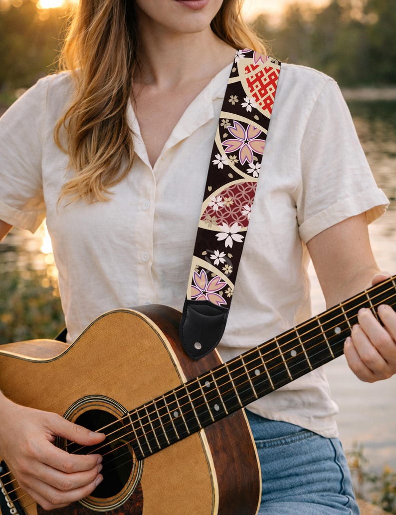 May include: A guitar strap with a black background and a Japanese-inspired floral design. The strap features pink cherry blossoms, white flowers, and red geometric patterns. The strap is attached to a wooden acoustic guitar.