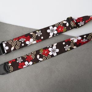 May include: A guitar strap with a dark brown background, adorned with a repeating pattern of white and red cherry blossoms and gold wave designs. The strap has black leather ends and a black adjustable buckle.