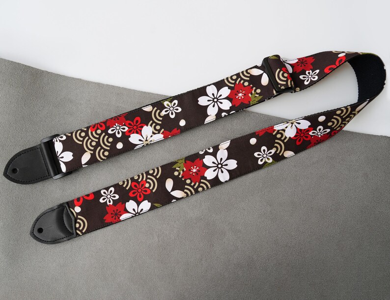 May include: A guitar strap with a dark brown background, adorned with a repeating pattern of white and red cherry blossoms and gold wave designs. The strap has black leather ends and a black adjustable buckle.