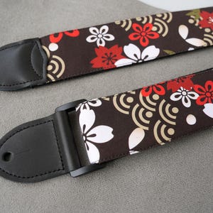 May include: Two guitar straps with a brown floral pattern featuring red and white flowers and a cream-colored wave design. The straps have black leather ends and a black adjustable buckle.