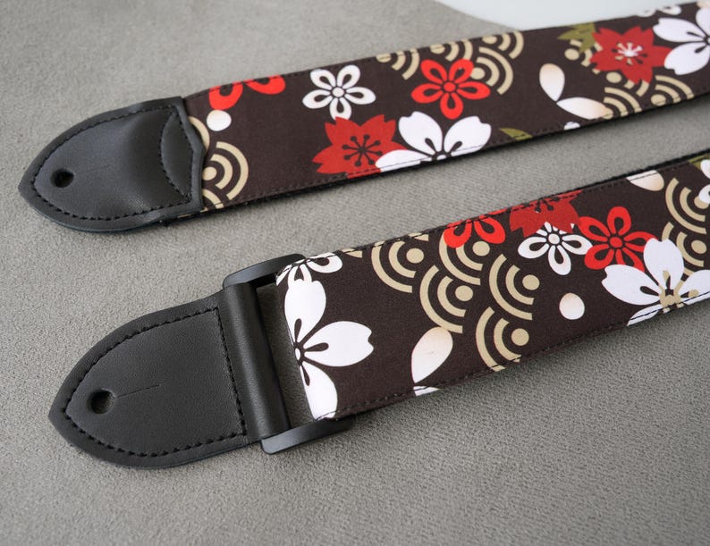 May include: Two guitar straps with a brown floral pattern featuring red and white flowers and a cream-colored wave design. The straps have black leather ends and a black adjustable buckle.