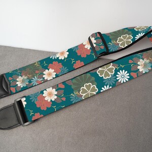 May include: A teal guitar strap with a floral pattern featuring red, white, and green flowers. The strap has black leather ends and a black adjustable buckle. The floral design is set against a teal background.