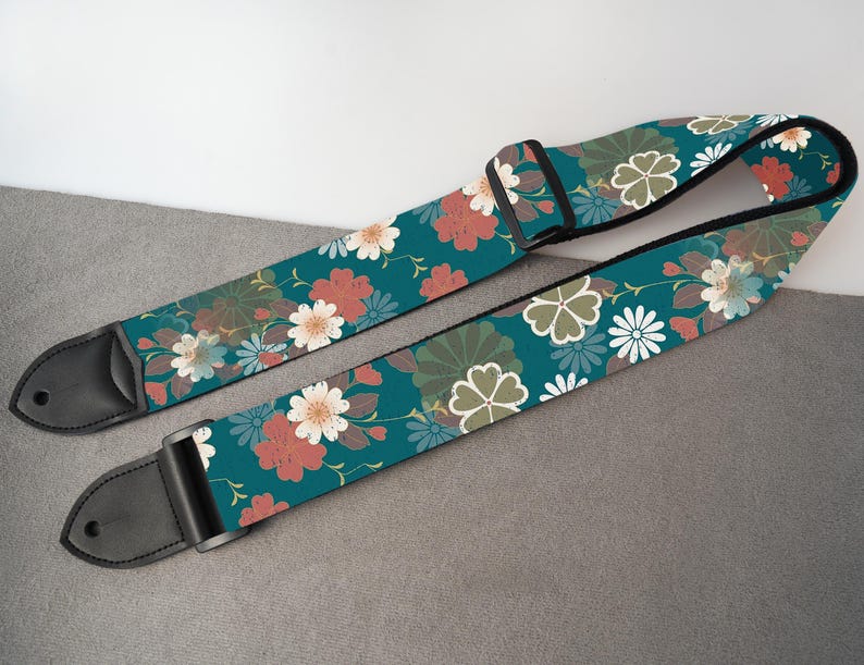 May include: A teal guitar strap with a floral pattern featuring red, white, and green flowers. The strap has black leather ends and a black adjustable buckle. The floral design is set against a teal background.