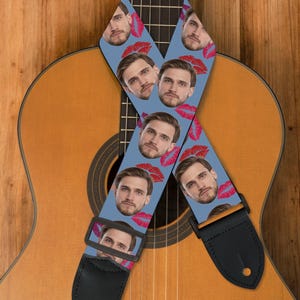 May include: A light blue guitar strap featuring a repeating pattern of a man's face and red lips. The strap is made of a durable material with black leather ends and a black adjustable buckle. The strap is resting on a wooden acoustic guitar.