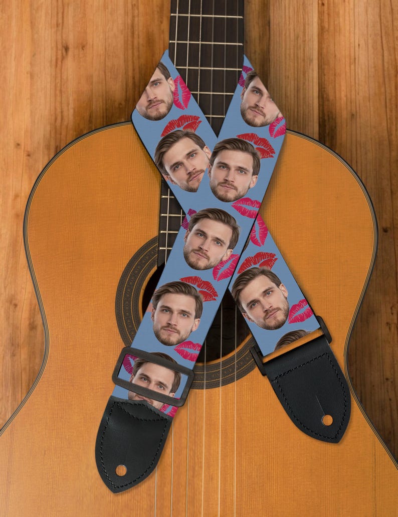 May include: A light blue guitar strap featuring a repeating pattern of a man's face and red lips. The strap is made of a durable material with black leather ends and a black adjustable buckle. The strap is resting on a wooden acoustic guitar.