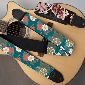 May include: Three guitar straps with floral patterns rest on an acoustic guitar. One strap features a teal background with red and white flowers. Another has a dark background with pink and white flowers. The third strap has a teal background with white and green floral designs.