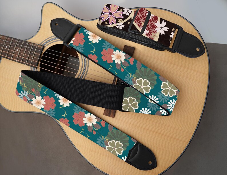 May include: Three guitar straps with floral patterns rest on an acoustic guitar. One strap features a teal background with red and white flowers. Another has a dark background with pink and white flowers. The third strap has a teal background with white and green floral designs.