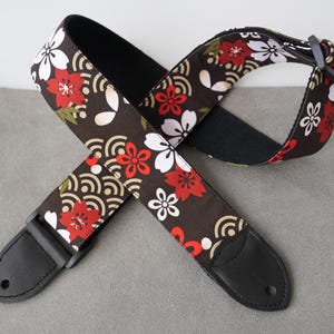 May include: A guitar strap with a floral pattern in red, white, and green on a dark brown background. The strap has black leather ends and a black adjustable buckle. The design features stylized flowers and wave patterns.