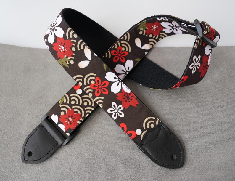 May include: A guitar strap with a floral pattern in red, white, and green on a dark brown background. The strap has black leather ends and a black adjustable buckle. The design features stylized flowers and wave patterns.