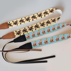 May include: Three camera straps with decorative patterns. One strap is light yellow with cat faces and paw prints, another is light blue with pig faces, and the third is light yellow with cat faces.