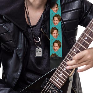 May include: A teal guitar strap featuring a repeating pattern of a person's face and musical notes. The strap is attached to a black electric guitar. The person is wearing a black leather jacket and a dog tag necklace.