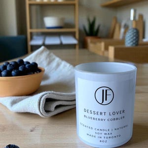 Op de afbeelding: Een witte geurkaars met de tekst "Dessert Lover Blueberry Cobbler" en "Made in Toronto 8oz". Een kleine oranje kom met bosbessen en een gevouwen witte doek staan op de achtergrond.