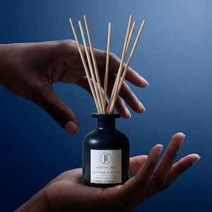 Puede incluir: Un difusor de caña azul oscuro con cañas de madera, sostenido en manos sobre un fondo azul. La etiqueta dice "MAISON BOIS LEATHER & WOOD" y "100ml 3.4 fl oz REED DIFFUSER".
