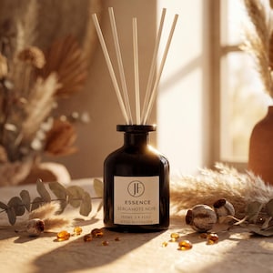 Könnte beinhalten: Eine dunkelbraune Glas-Duftflasche mit weißen Stäbchen. Das Etikett lautet "ESSENCE BERGAMOTE NOIR 100ml 3.4 fl oz REED DIFFUSER". Die Flasche ist von getrockneten Pflanzen und bernsteinfarbenen Ziersteinen umgeben.