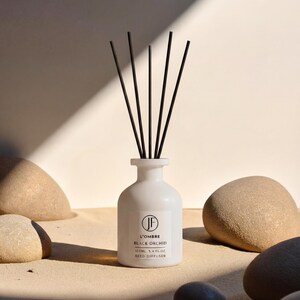 Op de afbeelding: Een witte rietverspreiderfles met zwarte rietjes, met het label "L'OMBRE BLACK ORCHID". De fles is omgeven door gladde, lichtbruine stenen. Het product is een geurartikel voor in huis.