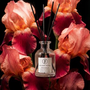 May include: A clear glass reed diffuser with black reeds, labeled "Bloom Petal Magnolia." The diffuser is surrounded by large, vibrant peach and burgundy iris flowers against a black background. The bottle contains 3.4 fl oz of liquid.