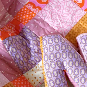 May include: A patchwork quilt with a variety of pink, orange, and purple fabrics. The quilt is made up of squares and rectangles of different sizes and patterns. Two purple pillows with a white floral pattern are in the foreground.