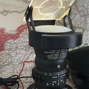 May include: A black reflector gun sight with a square, illuminated screen and a circular target reticle. The base has a dial for range and a small plaque that reads "SPITFIRE". The device is set against a map background.