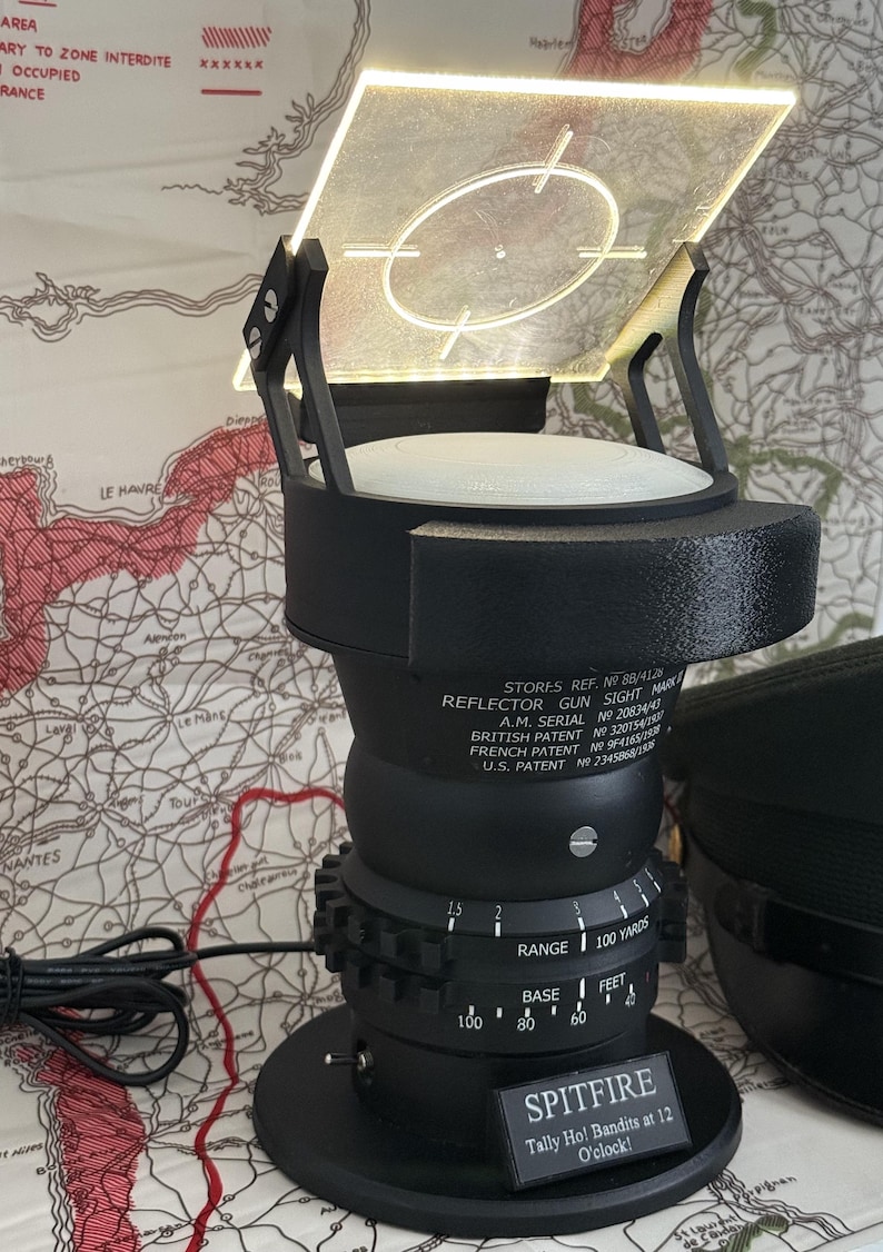 May include: A black reflector gun sight with a square, illuminated screen and a circular target reticle. The base has a dial for range and a small plaque that reads "SPITFIRE". The device is set against a map background.