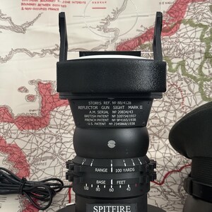 May include: Black reflector gun sight, Mark II, with a clear plastic aiming reticle. The sight has a circular base and a series of adjustable dials. The text "SPITFIRE" and "Tally Ho! Bandits at 12 O'clock!" are visible. The sight is on a map.