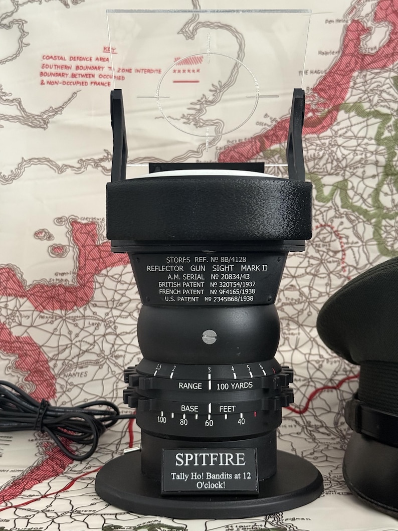 May include: Black reflector gun sight, Mark II, with a clear plastic aiming reticle. The sight has a circular base and a series of adjustable dials. The text "SPITFIRE" and "Tally Ho! Bandits at 12 O'clock!" are visible. The sight is on a map.