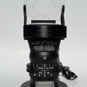 May include: A black reflector gun sight, Mark II, with a clear, rectangular lens and a circular reticle. The sight has a series of dials and the text "SPITFIRE Tally Ho! Bandits at 12 O'clock!" is visible. The base has a diameter of approximately 4 inches.