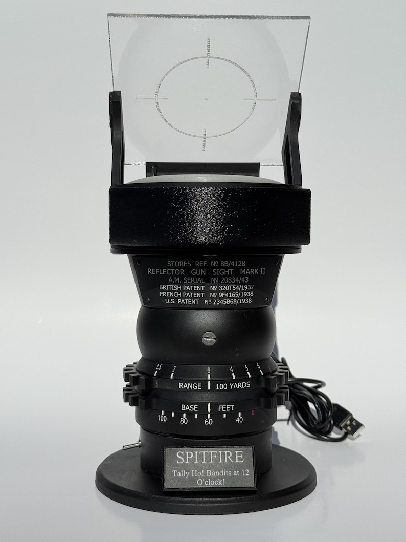 May include: A black reflector gun sight, Mark II, with a clear, rectangular lens and a circular reticle. The sight has a series of dials and the text "SPITFIRE Tally Ho! Bandits at 12 O'clock!" is visible. The base has a diameter of approximately 4 inches.