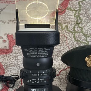 May include: A black reflector gun sight with a clear, illuminated screen featuring a crosshair target. The base reads "SPITFIRE" and "Tally Ho! Bandits at 12 O'clock!". A military-style hat is visible in the background.