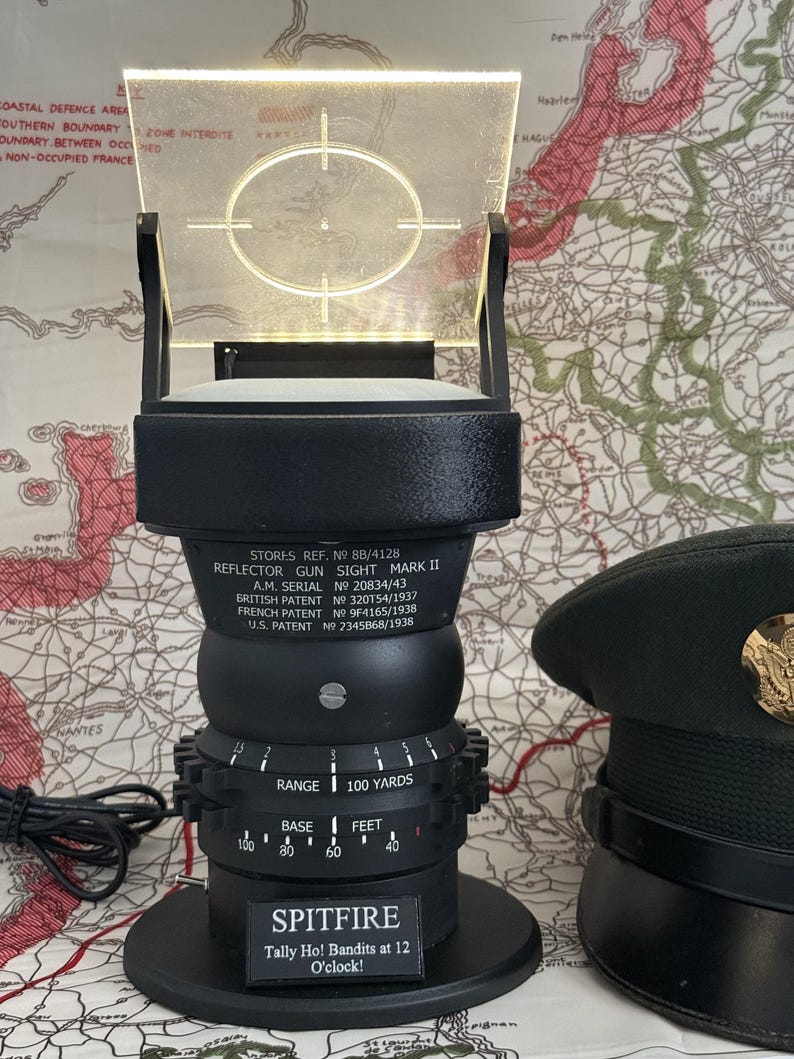May include: A black reflector gun sight with a clear, illuminated screen featuring a crosshair target. The base reads "SPITFIRE" and "Tally Ho! Bandits at 12 O'clock!". A military-style hat is visible in the background.
