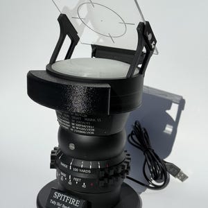 May include: A black and clear gun sight device with a clear, angled screen and a circular target design. The base has a label that reads "SPITFIRE" and "Tally Ho! Bandits at 12 O'clock!". The device has a USB cable attached.