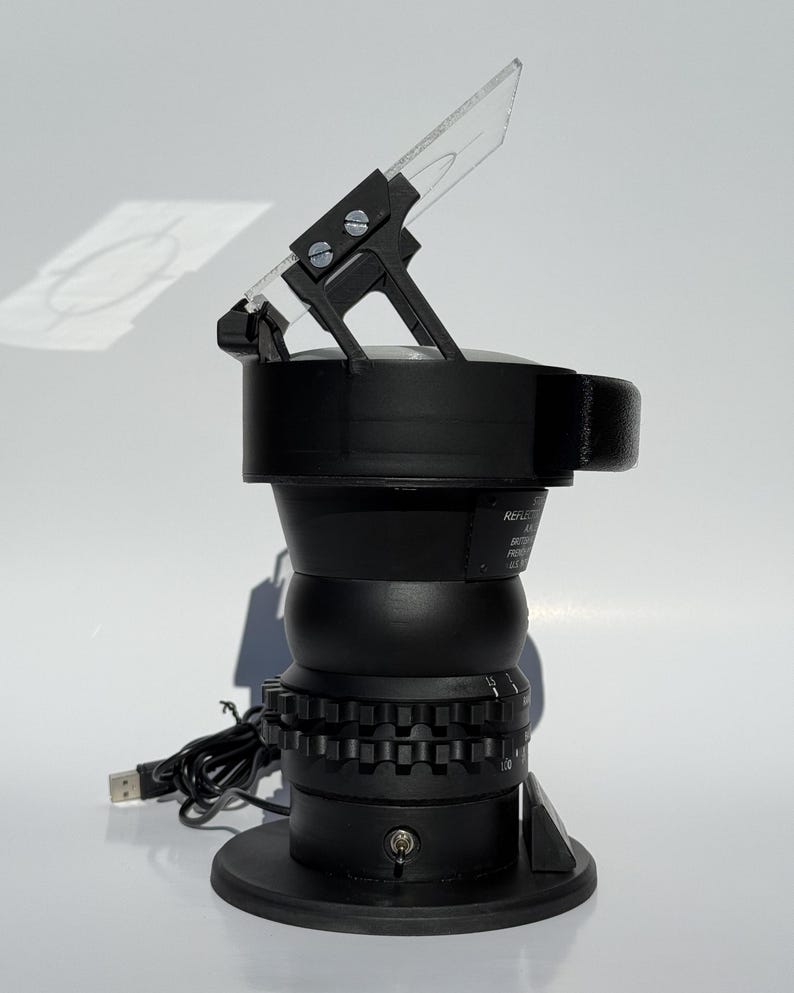 May include: Black optical instrument with a clear, angled lens. The device has a cylindrical body with a series of gears and a USB cable. The instrument is designed to reflect light, as indicated by the text on the side.