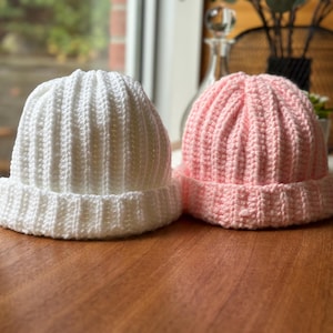 May include: Two crocheted hats, one white and one pink, on a wooden surface.