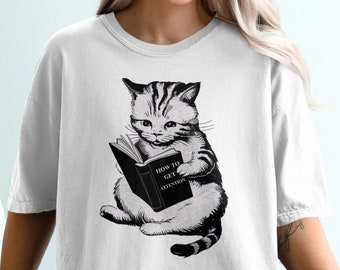 Könnte beinhalten: Weißes T-Shirt mit einer Schwarzweißillustration einer Katze, die ein Buch mit dem Titel "Wie man Aufmerksamkeit bekommt" liest.
