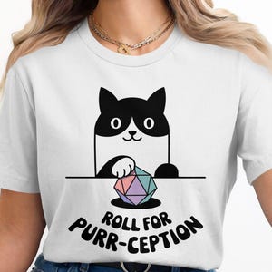 Pode incluir: Camiseta branca com uma ilustração em preto e branco de um gato segurando um dado multicolorido. O texto "Roll for Purr-ception" está impresso abaixo do gato.