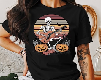 Peut inclure: Un t-shirt noir avec un graphique rétro d'un squelette jouant de la guitare avec deux citrouilles devant lui. Le graphique a un fond de coucher de soleil avec des couleurs orange, jaune et rouge.
