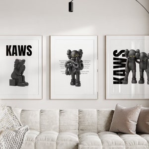May include: Three black and white framed prints with the word "KAWS" in bold black letters. The centre print features a black and white cartoon character holding a smaller cartoon character. The left print features a black and white cartoon character sitting with its head down. The right print features two black and white cartoon characters standing side by side.