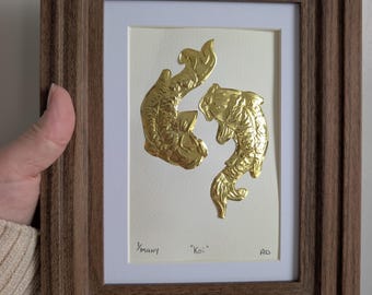 Puede incluir: Una obra de arte enmarcada con dos peces koi dorados. Los peces están en relieve sobre un fondo crema, con el texto "Koi" y "many" debajo. El marco es de madera marrón oscuro.