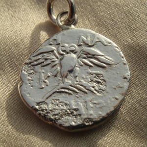 May include: A circular silver pendant featuring a relief of an owl with spread wings perched on a branch. The design includes the letters A, N, A, K, and S embossed on the surface. A small silver jump ring is attached to the top of the coin-shaped charm.