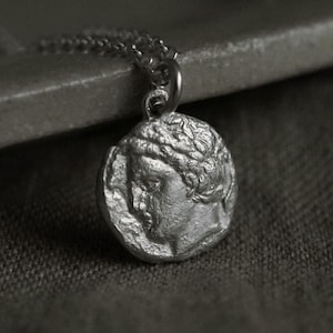 May include: Silver pendant necklace with a detailed relief of a classical figure's profile. The round pendant is suspended from a delicate chain. The design is reminiscent of ancient Greek or Roman art, suitable for everyday wear or special occasions.