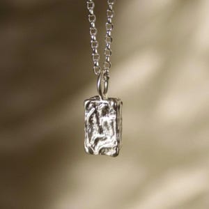 Peut inclure: Un collier en argent avec un pendentif rectangulaire texturé. Le pendentif a un aspect brut et organique.