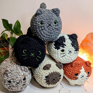 May include: Six crocheted cat plushies in various colors and patterns. The plushies are arranged in a pyramid shape, with the gray cat on top. The plushies have embroidered eyes and whiskers.