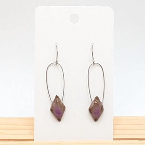 May include: A pair of dangle earrings featuring a diamond-shaped, purple and brown bead suspended from a silver-colored, elongated oval wire. The earrings are displayed on a white card against a light wood surface.