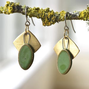 May include: A pair of handcrafted earrings featuring a brass square and a green oval pendant. The earrings hang from a simple hook. The earrings are displayed on a lichen-covered branch.