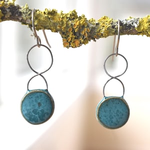 May include: A pair of handmade earrings with a unique design. Each earring features a silver-toned metal structure with a circular top, an infinity-shaped middle, and a round, turquoise-colored ceramic base. The earrings are displayed against a natural, lichen-covered branch.