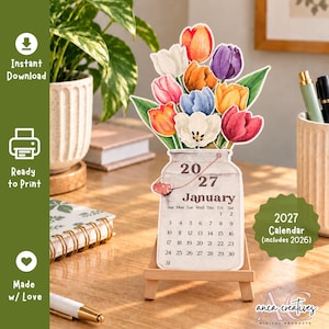 Può includere: Un calendario di gennaio 2027 stampato su un cartoncino fustellato a forma di barattolo di vetro riempito con un colorato bouquet di tulipani ad acquerello nei toni del rosso, arancione, giallo, bianco e viola, su un cavalletto in legno.