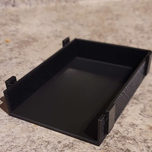 Puede incluir: Una caja de almacenamiento de plástico negro con una superficie texturizada. La caja está vacía y tiene una forma rectangular.