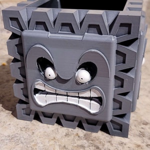 Op de afbeelding: Een grijze, 3D-geprinte Thwomp plantenbak uit de Super Mario-videogameserie. De plantenbak heeft een vierkante vorm met een gedetailleerd, boos gezicht en een getextureerde buitenkant. Hij is ontworpen om kleine planten of andere items in te bewaren.